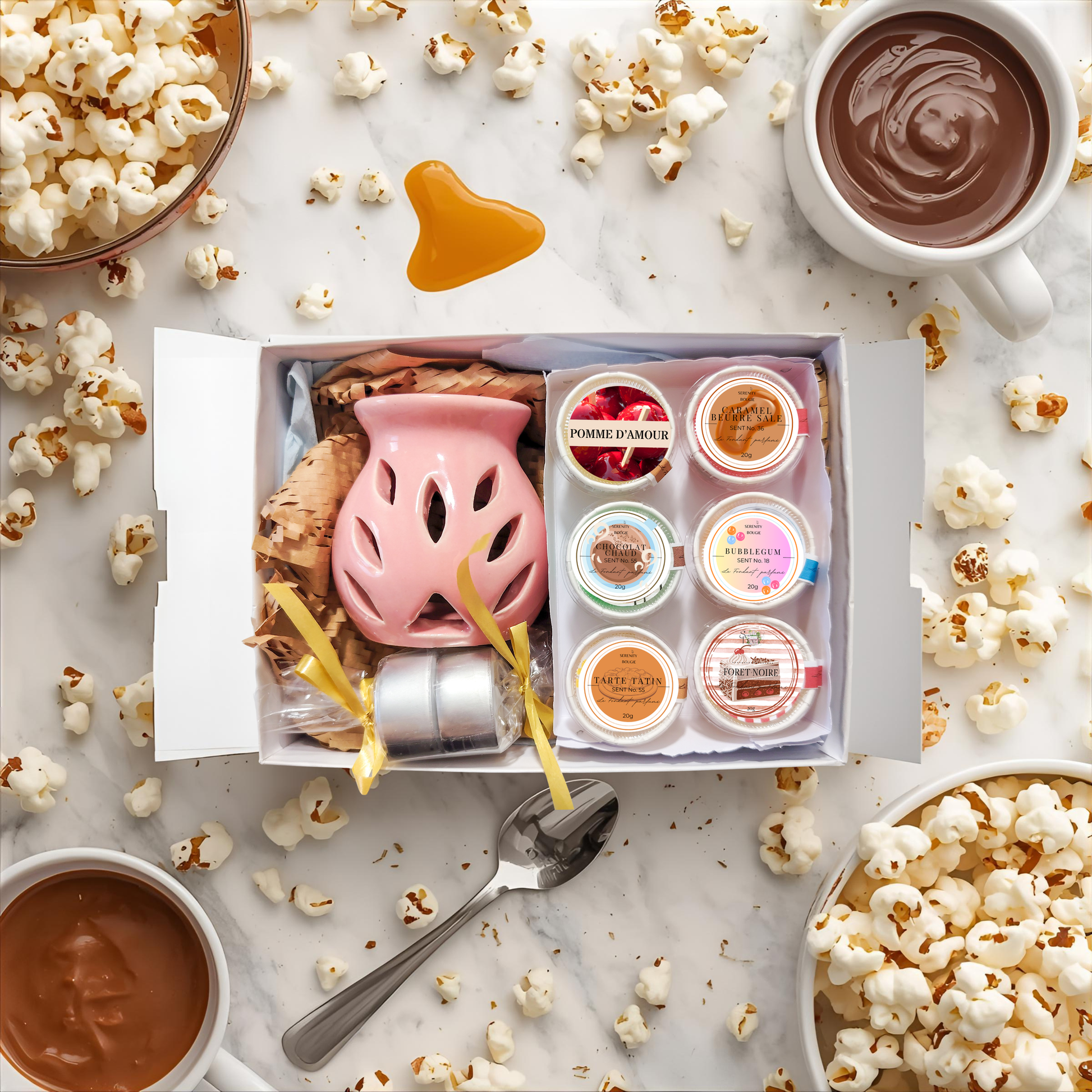 Coffret cadeau Fondants Parfumés Gourmands ouvert, contenant un brûle-parfum rose, 6 fondants (Caramel Beurre Salé, Chocolat Chaud, Pop Corn, Pomme d'Amour, Bubblegum, Tarte Tatin) et deux bougies. Décor de caramel coulant et de pop-corn.