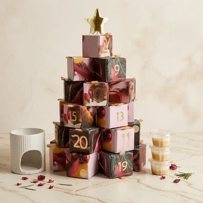 Une présentation de produit élégante sur une surface en marbre veiné blanc, montrant le calendrier de l'Avent Serenity Bougie. Le calendrier est un sapin formé de boîtes cadeaux décoratives numérotées, coiffé d'une étoile dorée. Un diffuseur d'huile en céramique blanche se trouve à gauche, et une pile de petits pots de bougies en cire est à droite. Des pétales de rose séchés et un brin de romarin sont délicatement dispersés sur le marbre. L'arrière-plan est un mur de marbre doux et flouté.