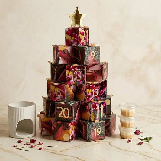 Une présentation de produit élégante sur une surface en marbre veiné blanc, montrant le calendrier de l'Avent Serenity Bougie. Le calendrier est un sapin formé de boîtes cadeaux décoratives numérotées, coiffé d'une étoile dorée. Un diffuseur d'huile en céramique blanche se trouve à gauche, et une pile de petits pots de bougies en cire est à droite. Des pétales de rose séchés et un brin de romarin sont délicatement dispersés sur le marbre. L'arrière-plan est un mur de marbre doux et flouté.