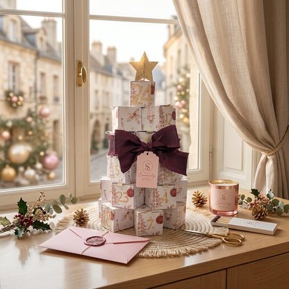 Une présentation de produit élégante sur une surface en bois montrant le calendrier de l'Avent Serenity Bougie. Le calendrier est un sapin formé de boîtes cadeaux décoratives numérotées, coiffé d'une étoile dorée. Un diffuseur d'huile en céramique blanche se trouve à gauche, et une pile de petits pots de bougies en cire est à droite. Des pétales de rose séchés et un brin de romarin sont délicatement dispersés sur le marbre. L'arrière-plan est une vue sur l'extérieur 