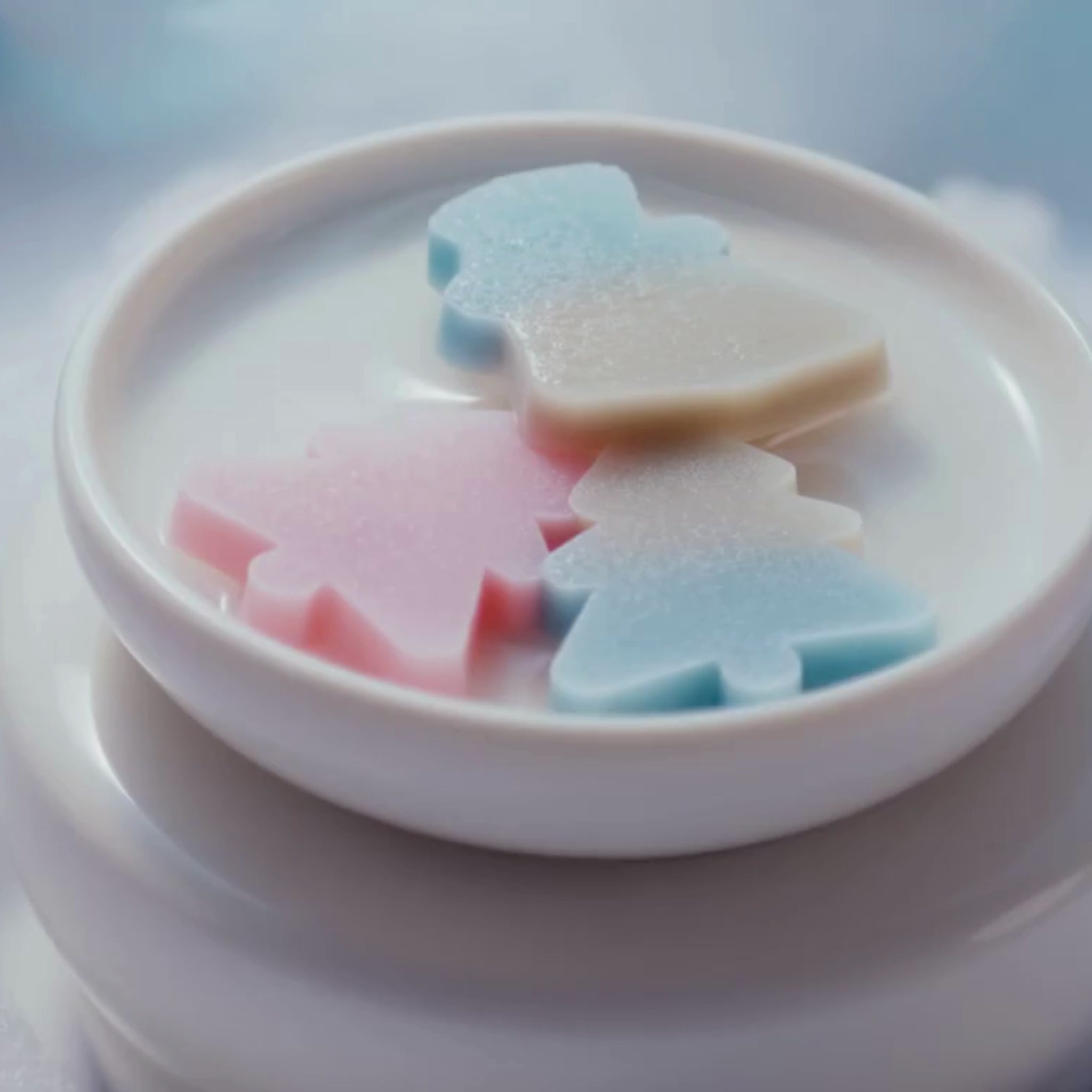 Vidéo en gros plan de trois fondants de cire Serenity Bougie en forme de mini sapins et bonhomme de neige aux couleurs pastel, scintillant avec du mica sur un brûleur blanc.