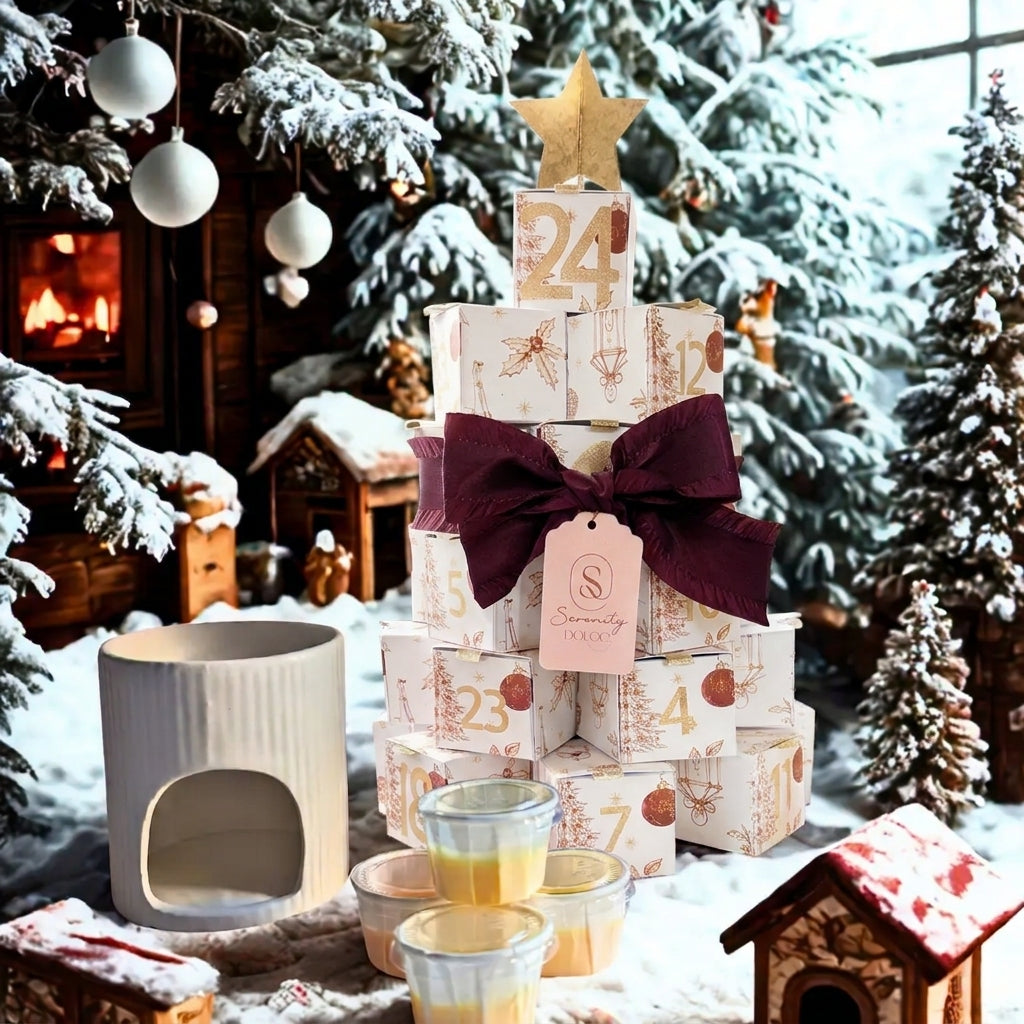 Une scène de Noël enchantée et enneigée mettant en scène le calendrier de l'Avent Serenity Bougie. Le calendrier est construit en forme de sapin de Noël avec des boîtes cadeaux numérotées et colorées (rose, rouge, noir), coiffé d'une étoile dorée. À sa base se trouvent un diffuseur d'huile en céramique blanche et une pile de petits pots de bougies en cire empilés. L'arrière-plan comprend des sapins enneigés décorés de boules blanches, une cheminée allumée et des maisons miniatures de Noël, créant une atmosp